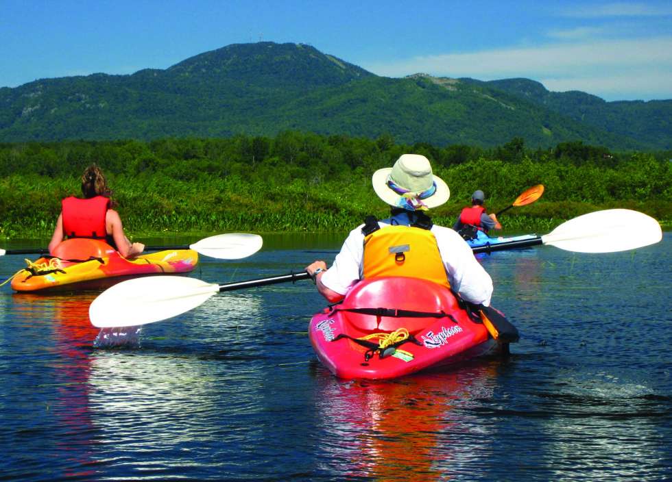 Trois personnes de dos chacun sur des kayaks colorés. Au loin, de la verdure et le Mont Orford