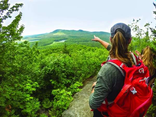 Femme de dos avec un sac à dos rouge qui pointe vers l'horizon au sommet d'une montagne en été.