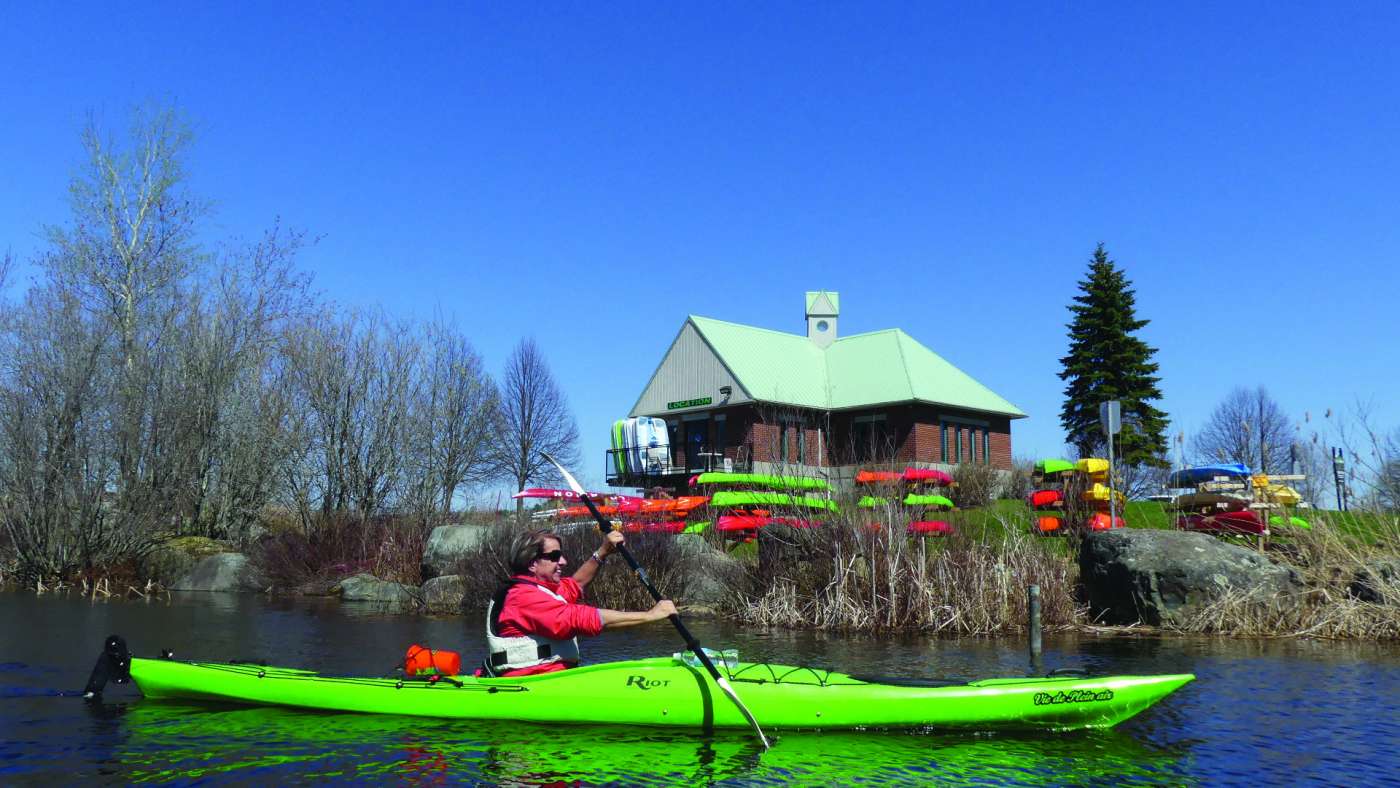 Homme à bord d'un kayak de mer vert lime sur le lac Memphrémagog. Derrière lui, le centre de location d'équipement de plein air, Vie de plein air.
