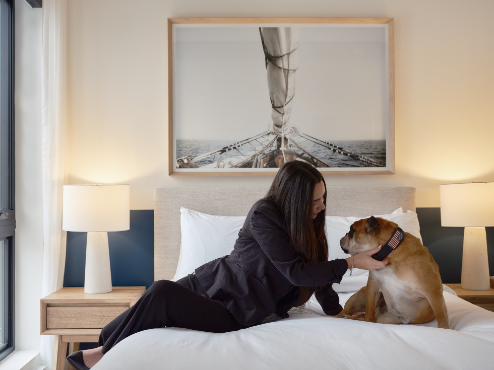 Une femme et son chien sont assis sur le lit de l'une des chambres de l'Hôtel Quartier des marinas de Magog